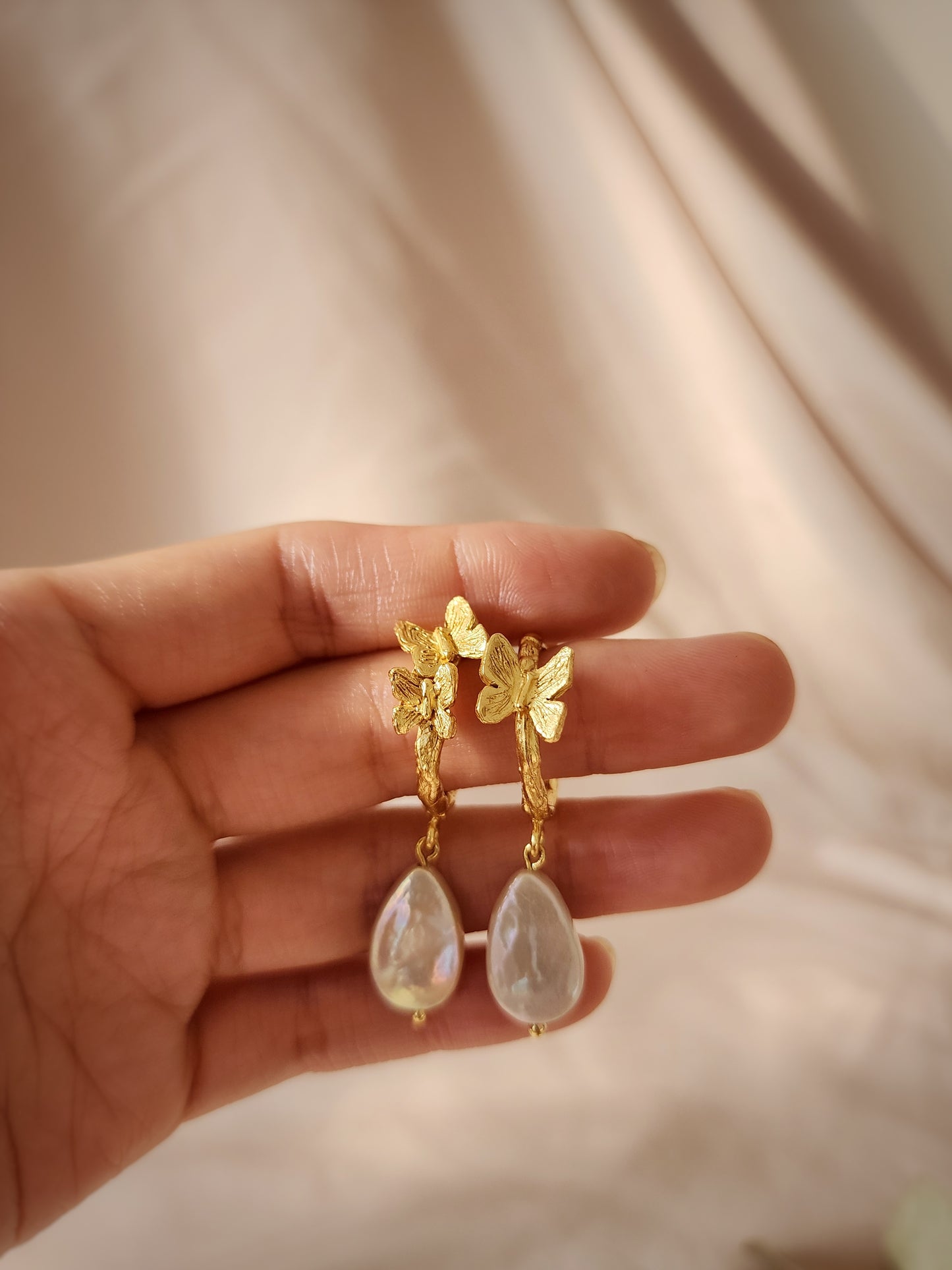 Hand sculpted pearl butterfly earrings in 18k gold vermeil held in hand to show scale, featuring intricate lost wax details and a warm studio glow.
