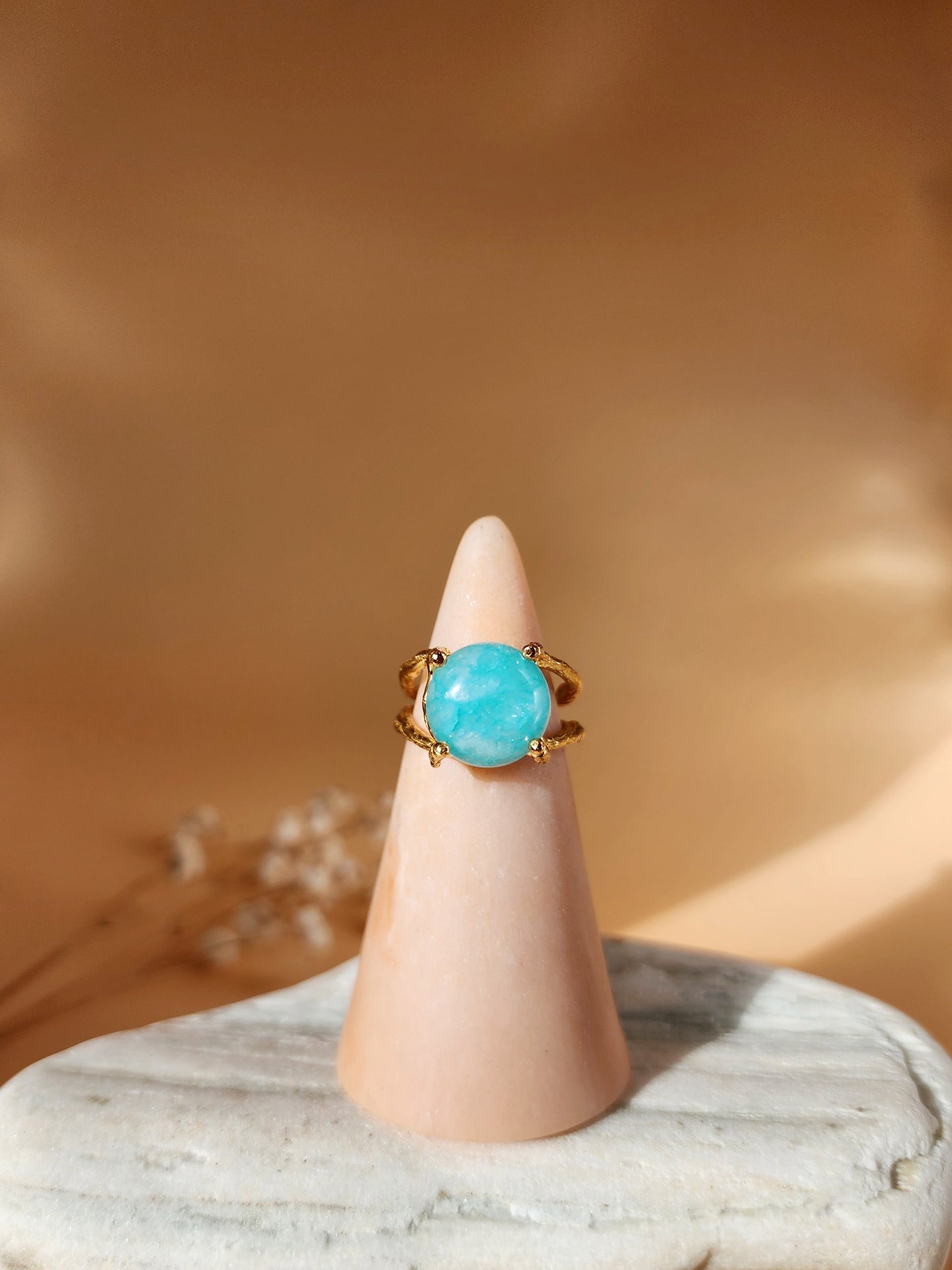 A hand sculpted gold ring featuring a natural 12 mm round Amazonite gemstone, displayed on a ring holder sitting on a white stone. The ring is set in a double gallery basket setting with organic details on the band, resembling natural branch like textures.