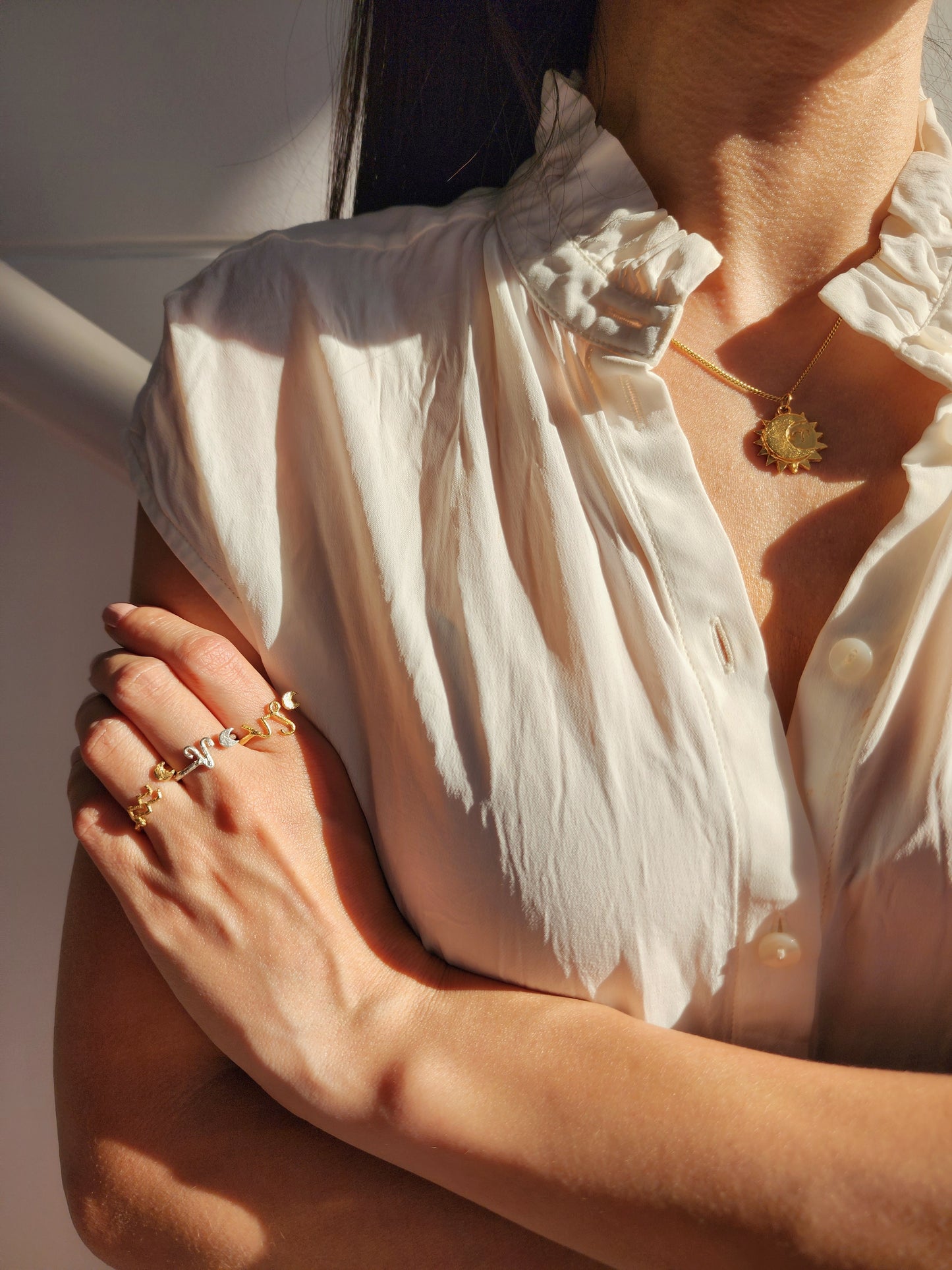 Three Astrology sign rings made of sterling silver and fine gold plated brass worn by a woman wearing a white short sleeve blouse with her arms are gently crossed, showcasing the handmade rings in warm sunlight.