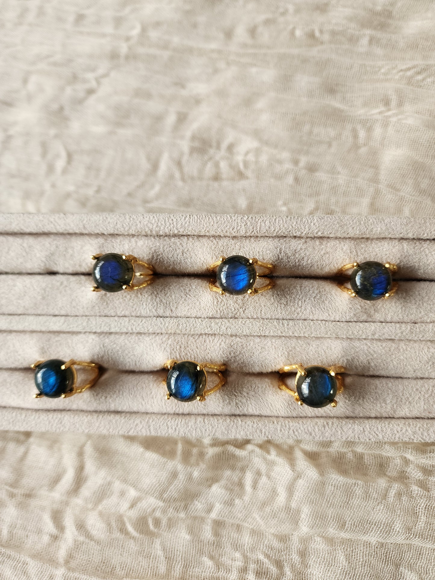 Six natural labradorite gemstone rings, each showing a different stone pattern and colour variation, displayed together in a white velvet jewellery tray.