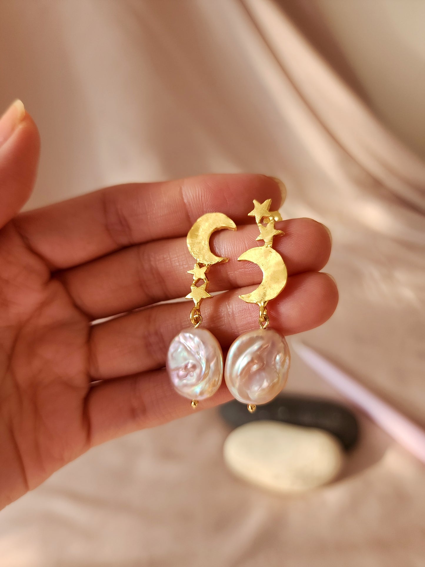 Celestial moon and star earrings in 18k gold vermeil, hand-sculpted in Sydney. A pink natural pearl dangles from the molten textured metal, illuminated by sunlight against an atmospheric backdrop of candles and stones to reflect our raw, organic aesthetic.