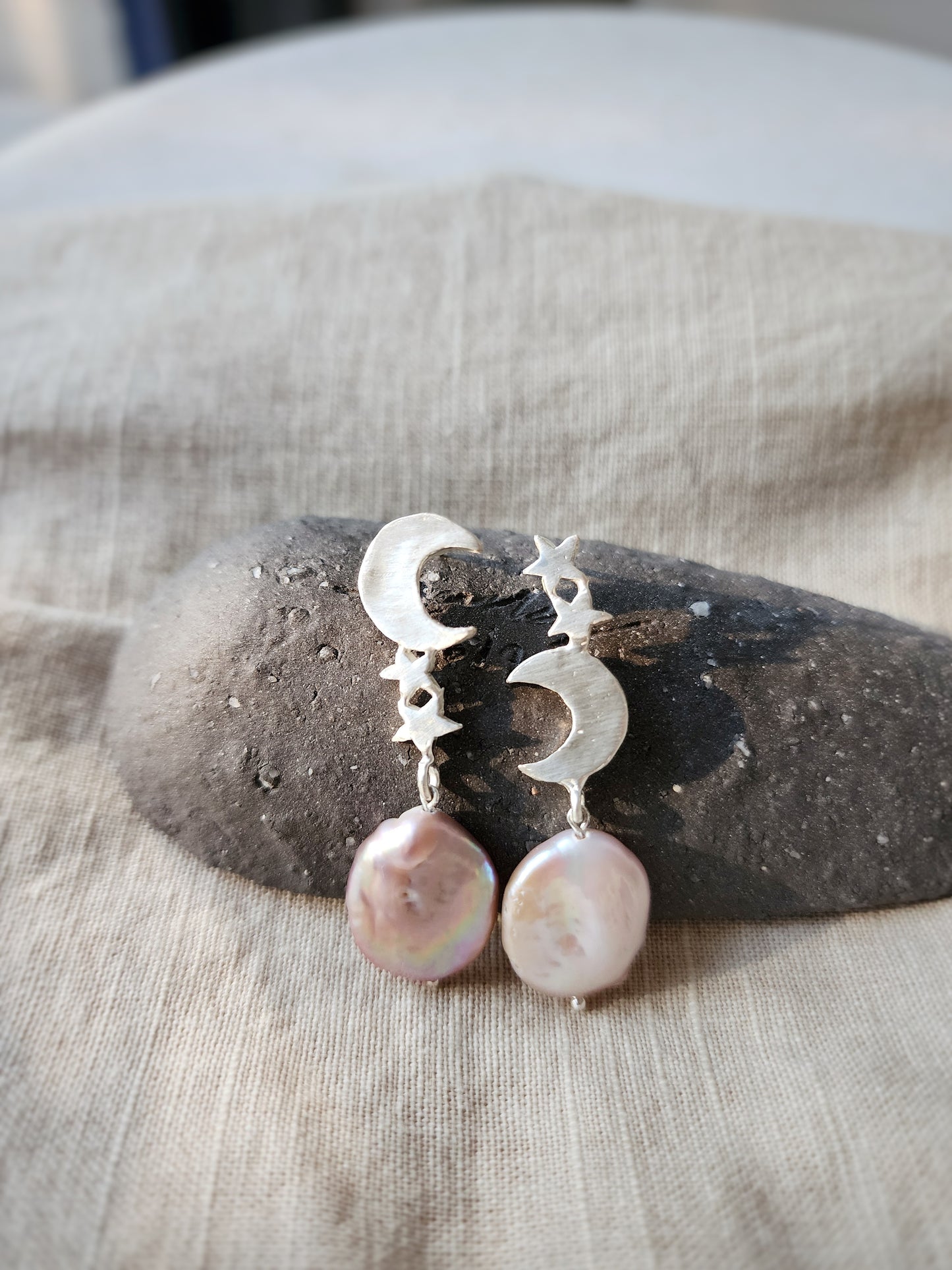 A pair of solid sterling silver astrology inspired earrings with pink pearls is styled on a grey rocky display stand on a soft linen cloth in our Sydney studio. The sunlight illuminates the metal, showcasing the intricate detail of the artisanal lost wax craft. 