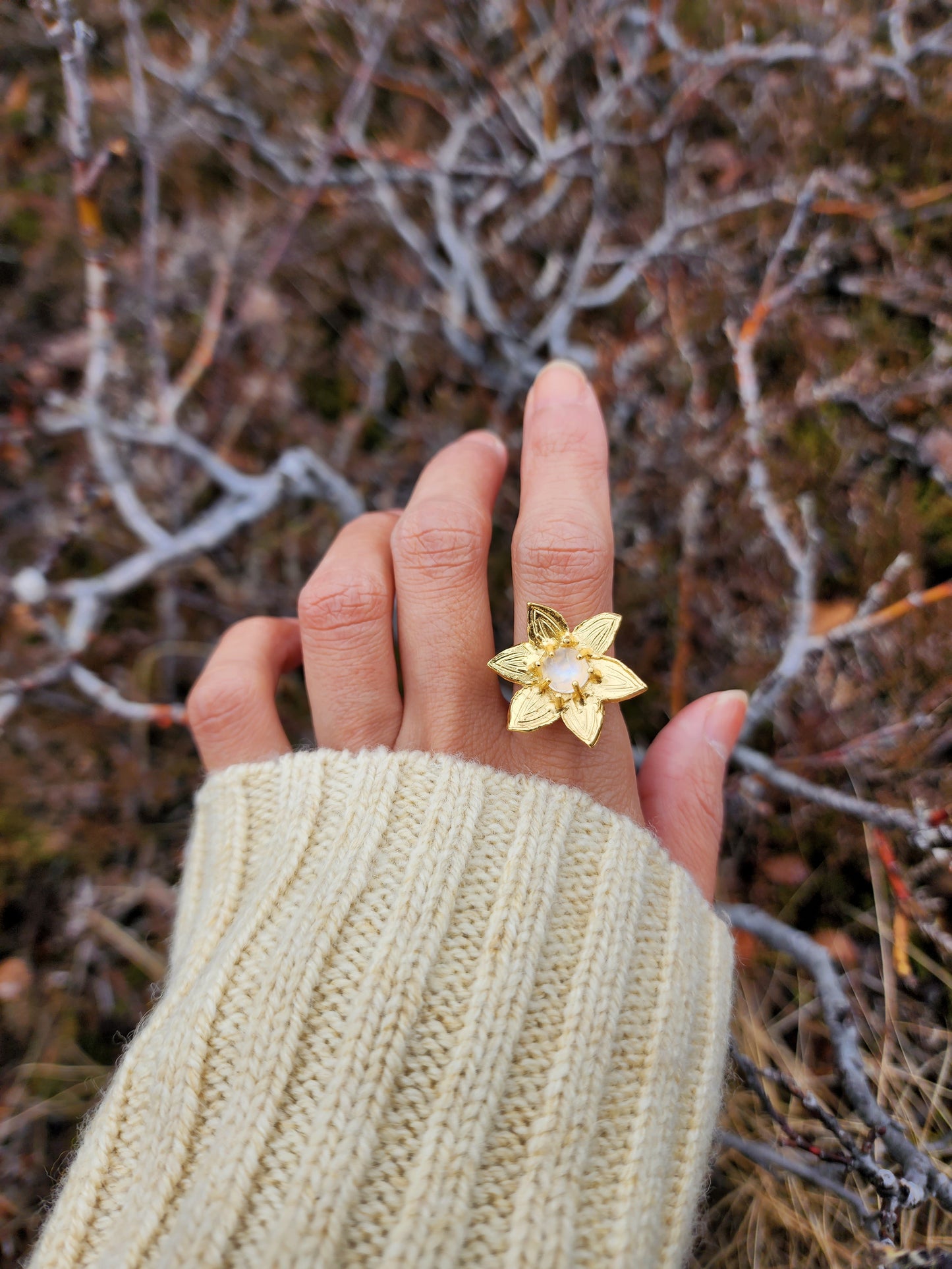 Moonstone flower ring featuring 7mm natural moonstone crystal. Worn on models hand to highlight size and fit and styled with yellow sweater. Shot in Iceland with wild looking branches in the background.