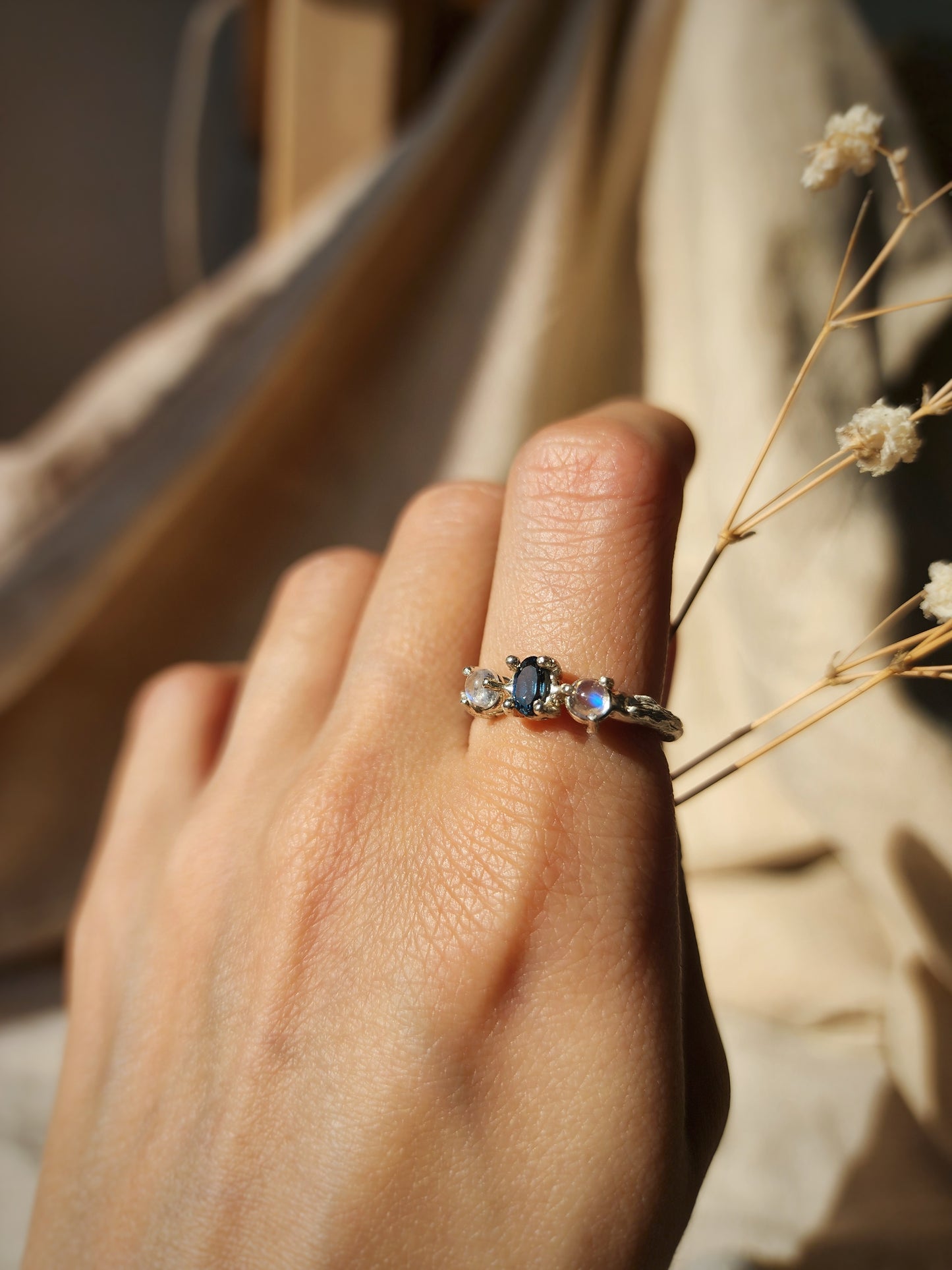 A Moonstone and blue topaz silver ring worn on hand to show scale and size. Styled holding dried flowers to highlight the natural inspiration behind the piece. 