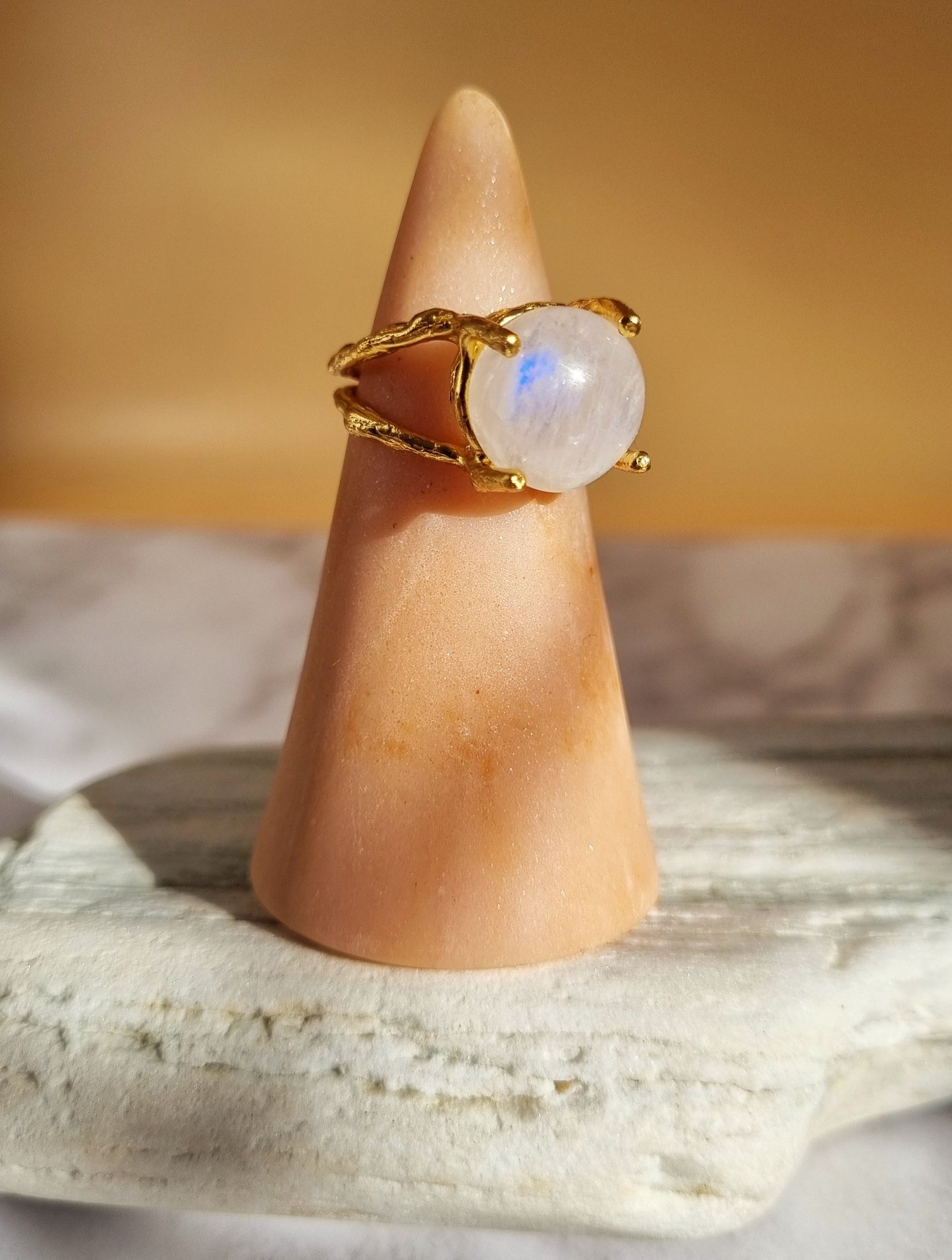 Side profile of a moonstone ring, hand sculpted and cast in recycled brass with fine 20k gold plating. The organic textured ring band captures a round 12mm moonstone in a four prong double gallery setting. Styled on a white stone. 