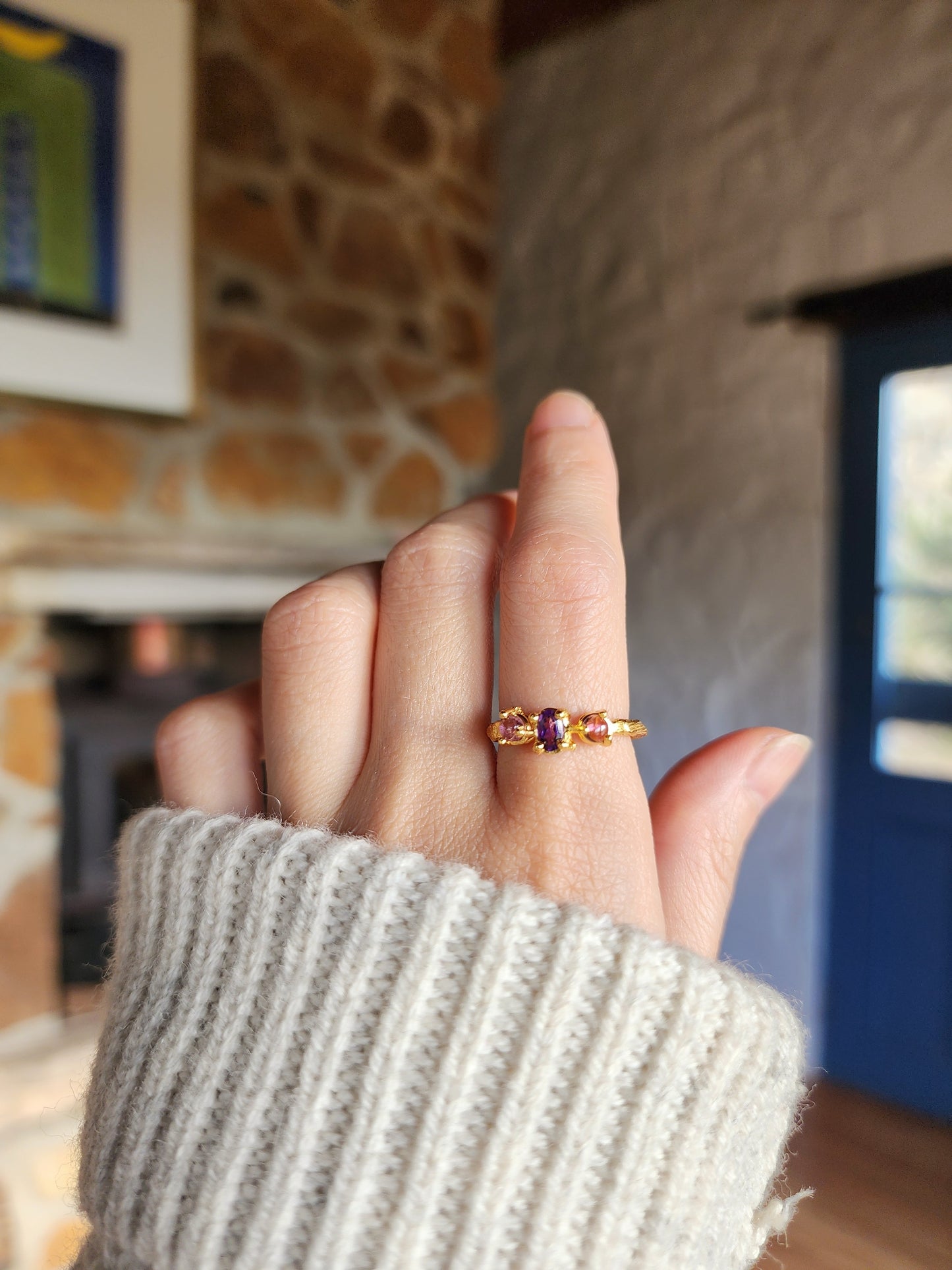 Triple gemstone ring featuring natural amethyst and pink tourmaline gemstones styled with a white wool sweater in an indoor cottage setting. 