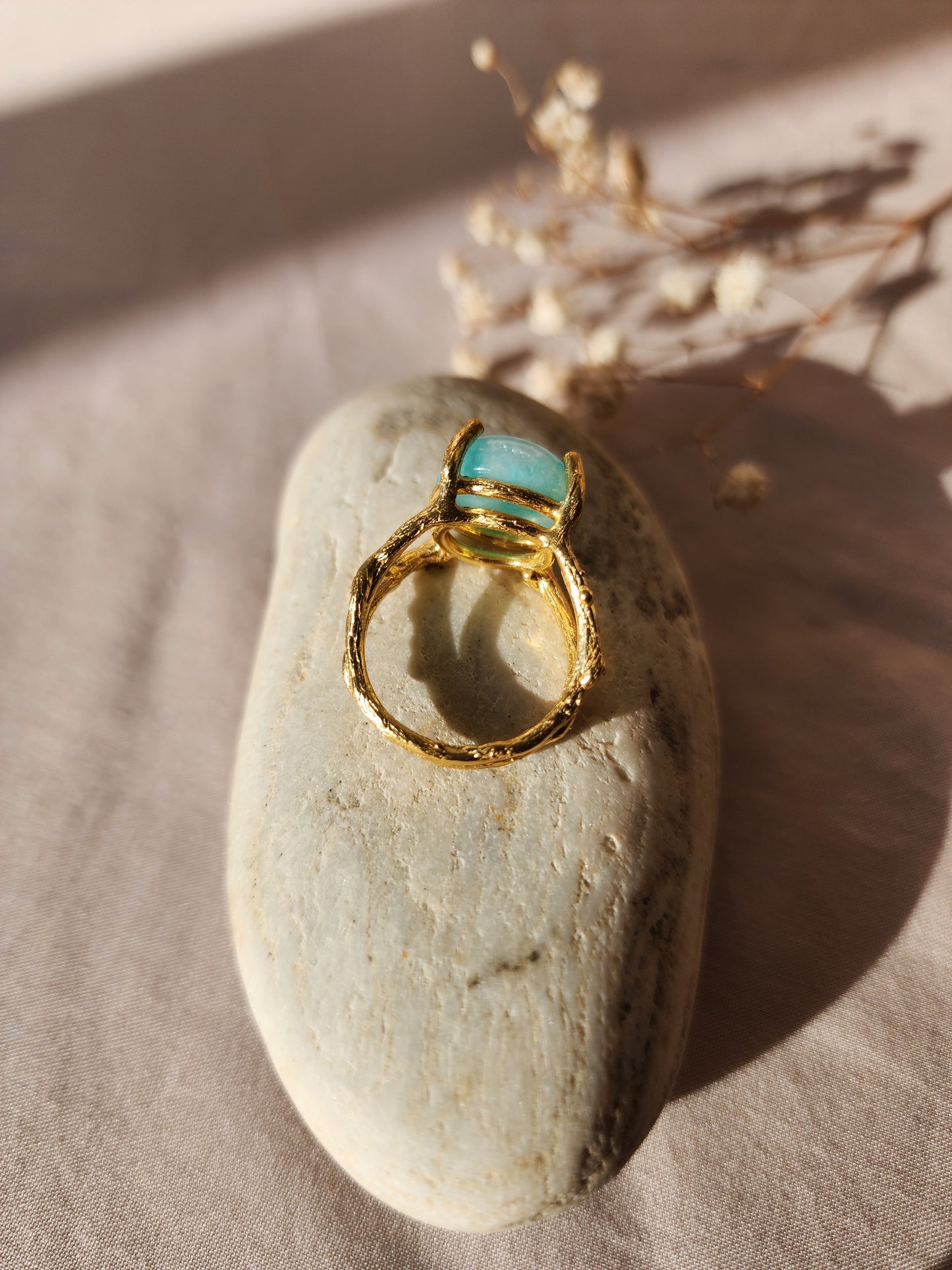 A top-down shot of a handmade 12mm natural Amazonite ring in a hand-sculpted gold double gallery setting. Crafted in our Sydney studio, the ring’s branch-like textures are displayed against a backdrop of dried flowers and white stone, emphasizing its artisanal, nature inspired origin.
