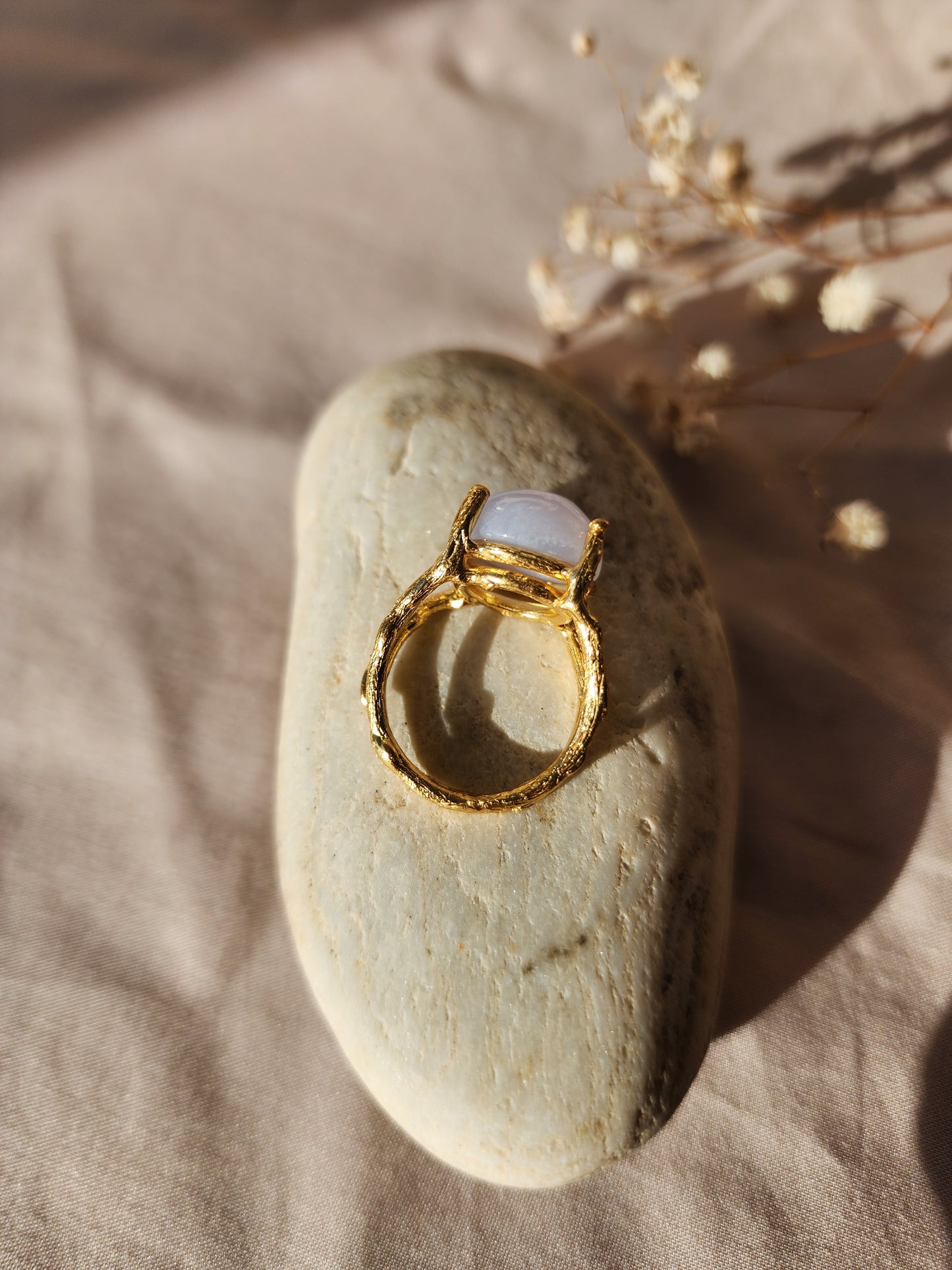A top down shot of a handmade natural blue lace agate ring in a hand-sculpted gold double gallery setting. Crafted in our Sydney studio, the ring’s branch-like organic textures are displayed against a backdrop of dried flowers and white stone, emphasizing its artisanal, nature inspired origin.