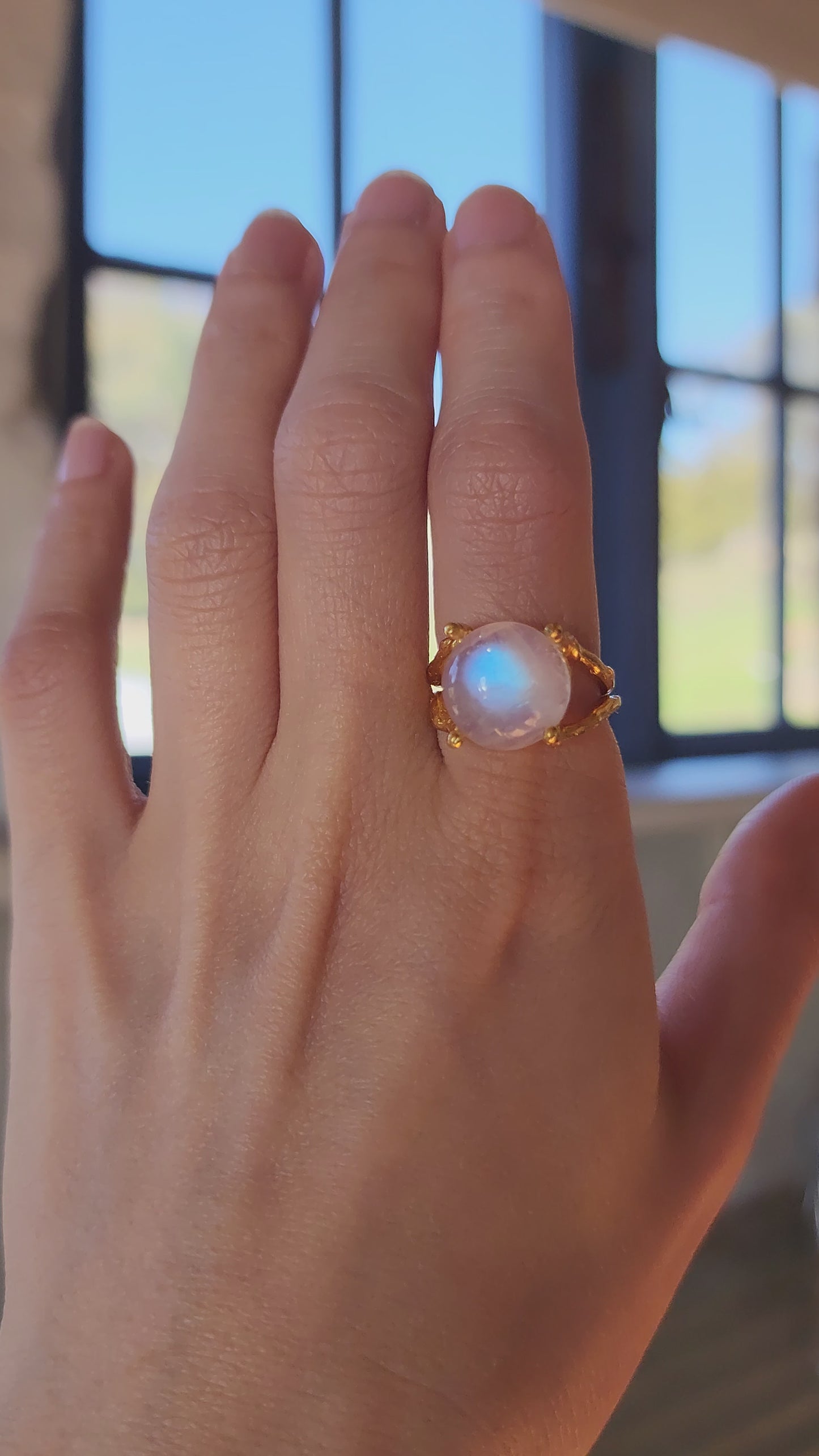 Woman's hand wearing a gold natural moonstone ring, the strong blue shine is highlighted as the hand moves closer to the camera. Worn in a Tasmanian cottage to highlight the Australian brand. 